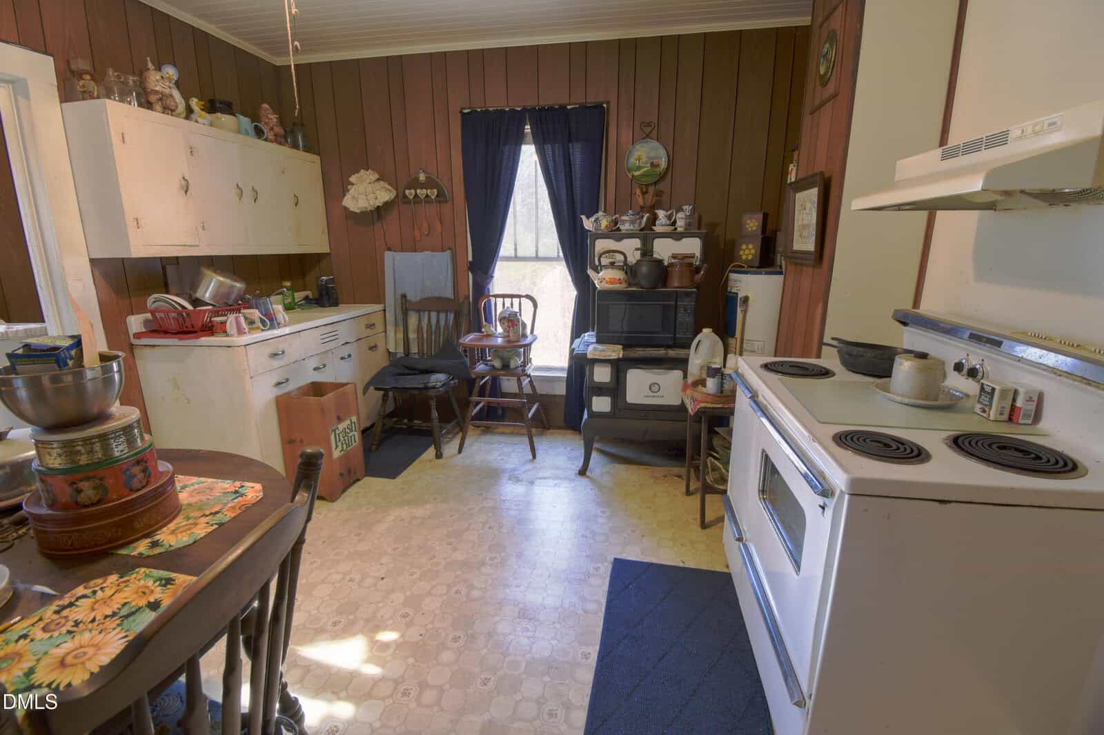 A vintage kitchen with wood-paneled walls, white cabinets, and an old-fashioned stove. A table with dishes and a bowl is on the left, while a stove and shelves with kitchen items are on the right. Sunlight shines through a window.