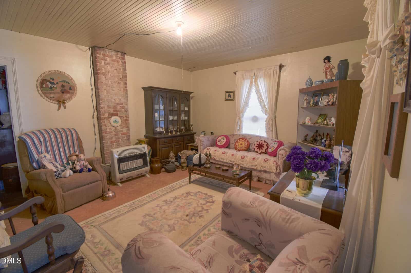 A cozy vintage living room with floral upholstered furniture, a brick-accented wall, a china cabinet, shelves with figurines, and a window with lacy curtains. Dolls and decorative items add a nostalgic touch.