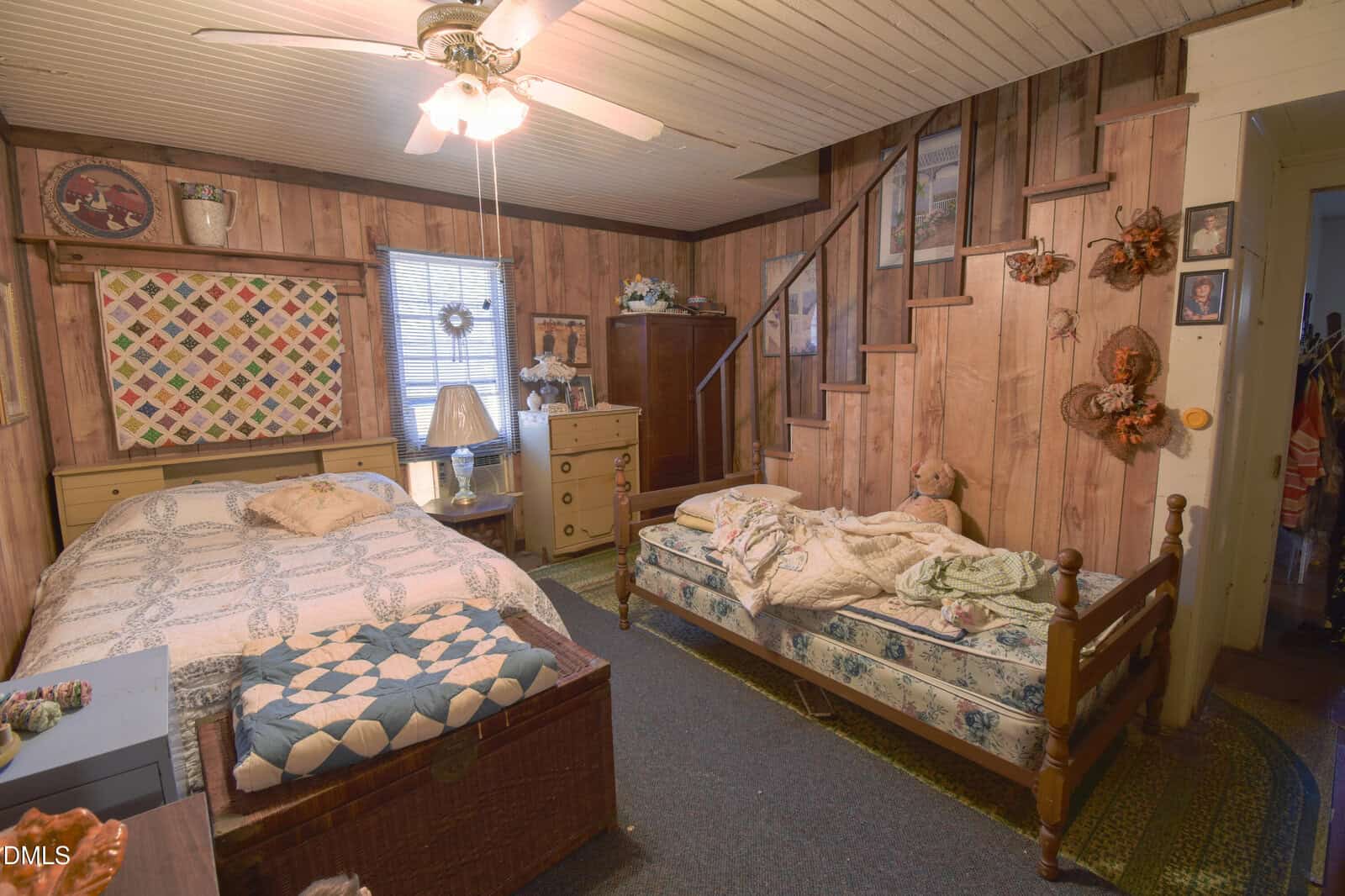 A cozy rustic bedroom with wood-paneled walls, two beds with patchwork quilts, a small dresser, wall decor, a ceiling fan, and stairs leading up. A teddy bear sits on one bed, and light streams in through a window.