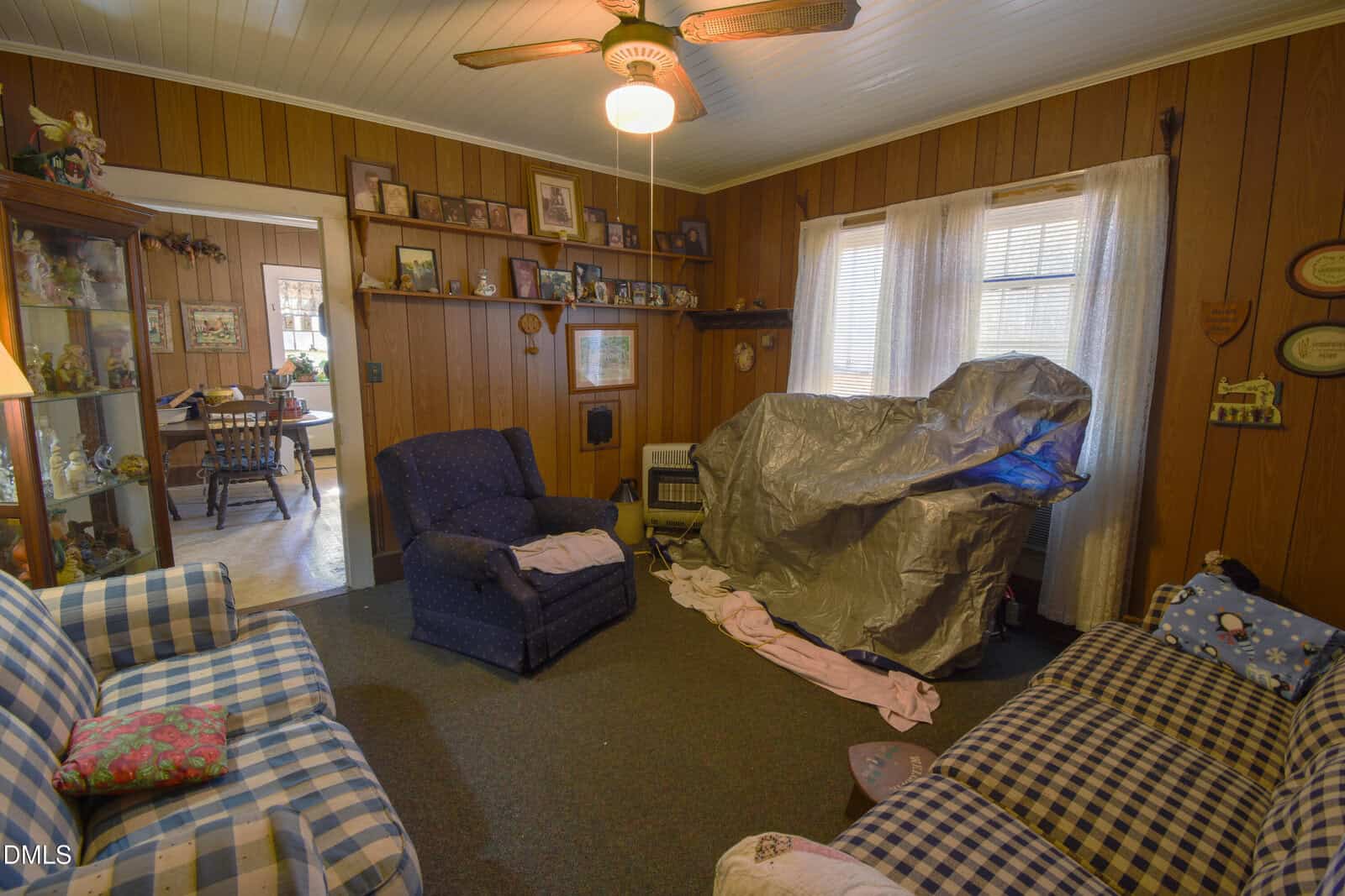 A cozy living room with wood-paneled walls, several armchairs and sofas, family photos on shelves, a ceiling fan, and a large object covered by a tarp near the window. A dining area is visible through an open doorway.