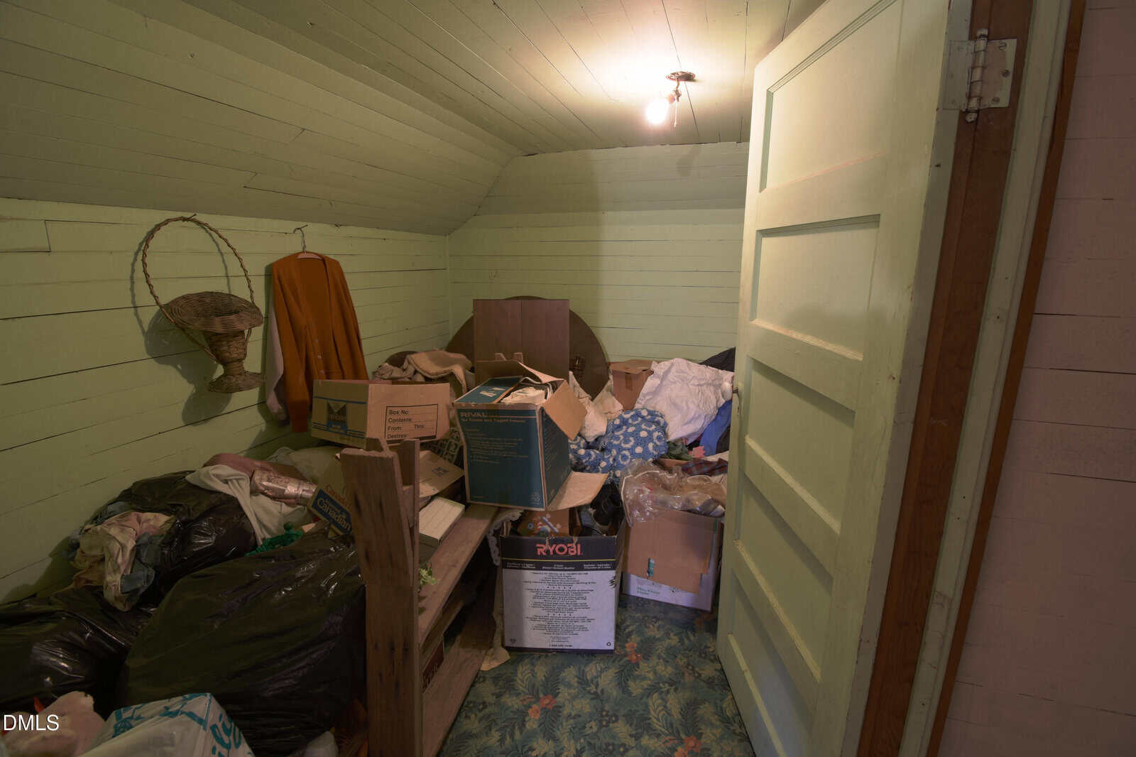 A small, cluttered room with green walls, an open door, boxes, bags, clothes, and miscellaneous items scattered on the floral carpet. A light bulb hangs from the ceiling.