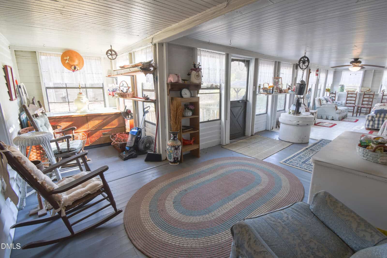 A bright, cozy sunroom with wooden floors, rocking chairs, a circular braided rug, shelves with decorations, and large windows letting in natural light. The room has country-style decor and overlooks an outdoor area.
