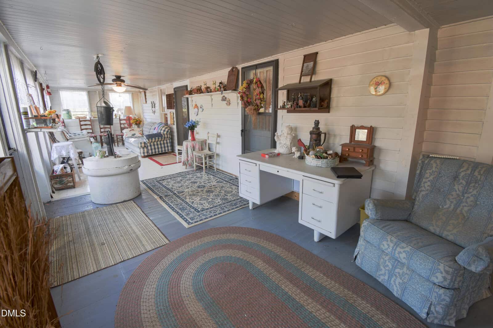 A cozy enclosed porch with wooden floors, area rugs, a white desk, blue armchair, various decorative items, and vintage furniture. The space is bright with natural light from windows along one wall.