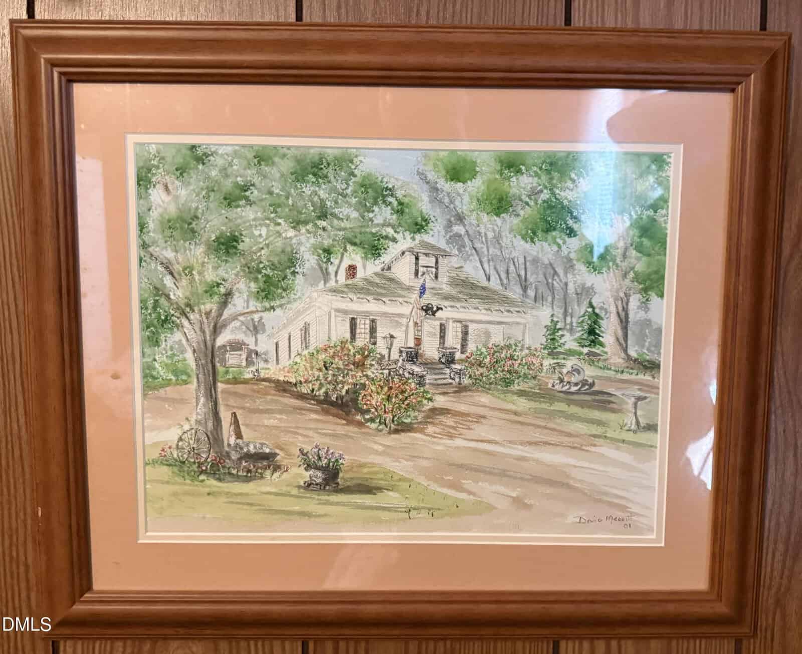A framed watercolor painting of a country house with a porch, surrounded by trees, colorful flowers, a birdbath, and garden decorations, hanging on a wood-paneled wall.