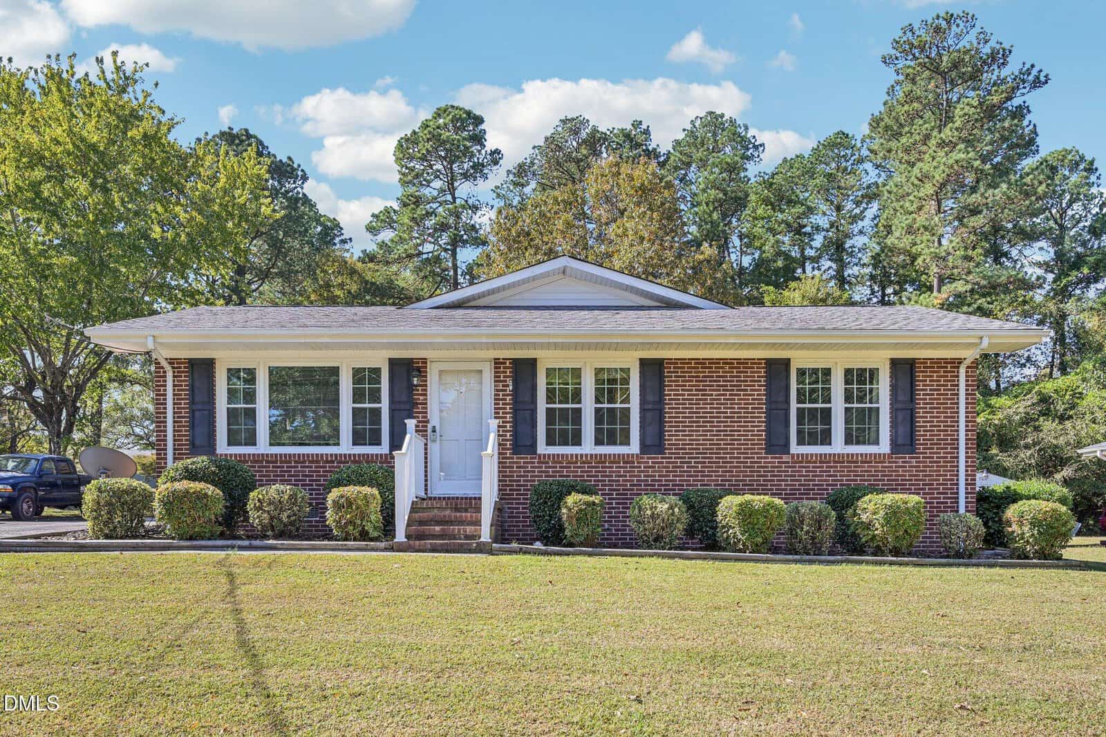 This charming one-story brick house on 309 20th Street in Butner features black shutters, white trim, and a cozy front porch with railings. It sits on a grassy lawn with neatly trimmed bushes and trees in the background.