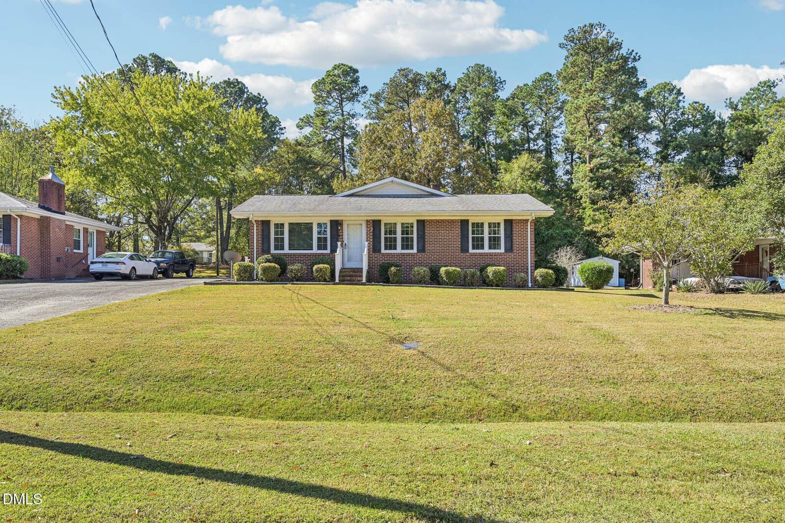 Single-story brick house with a covered front porch and white trim, set on a large lawn with neatly trimmed bushes. Trees and neighboring houses are visible in the background under a partly cloudy sky.