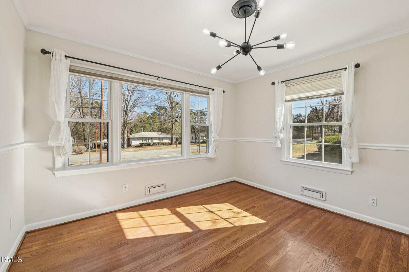 A bright, empty room with light-colored walls, two large windows with white curtains, hardwood flooring, and a modern black ceiling light fixture. Sunlight streams in, casting shadows on the floor.