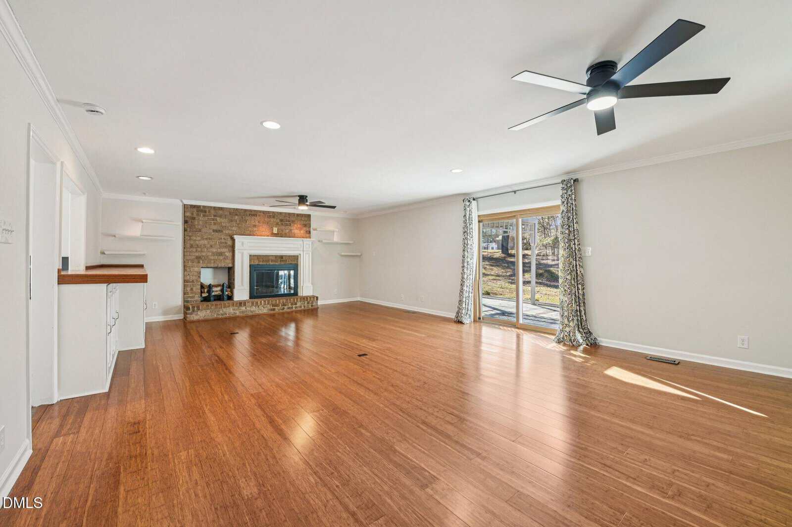 Spacious living room with wood flooring, brick fireplace, ceiling fans, built-in shelves, and sliding glass doors leading to a backyard patio. Room features neutral walls and natural light from large windows.