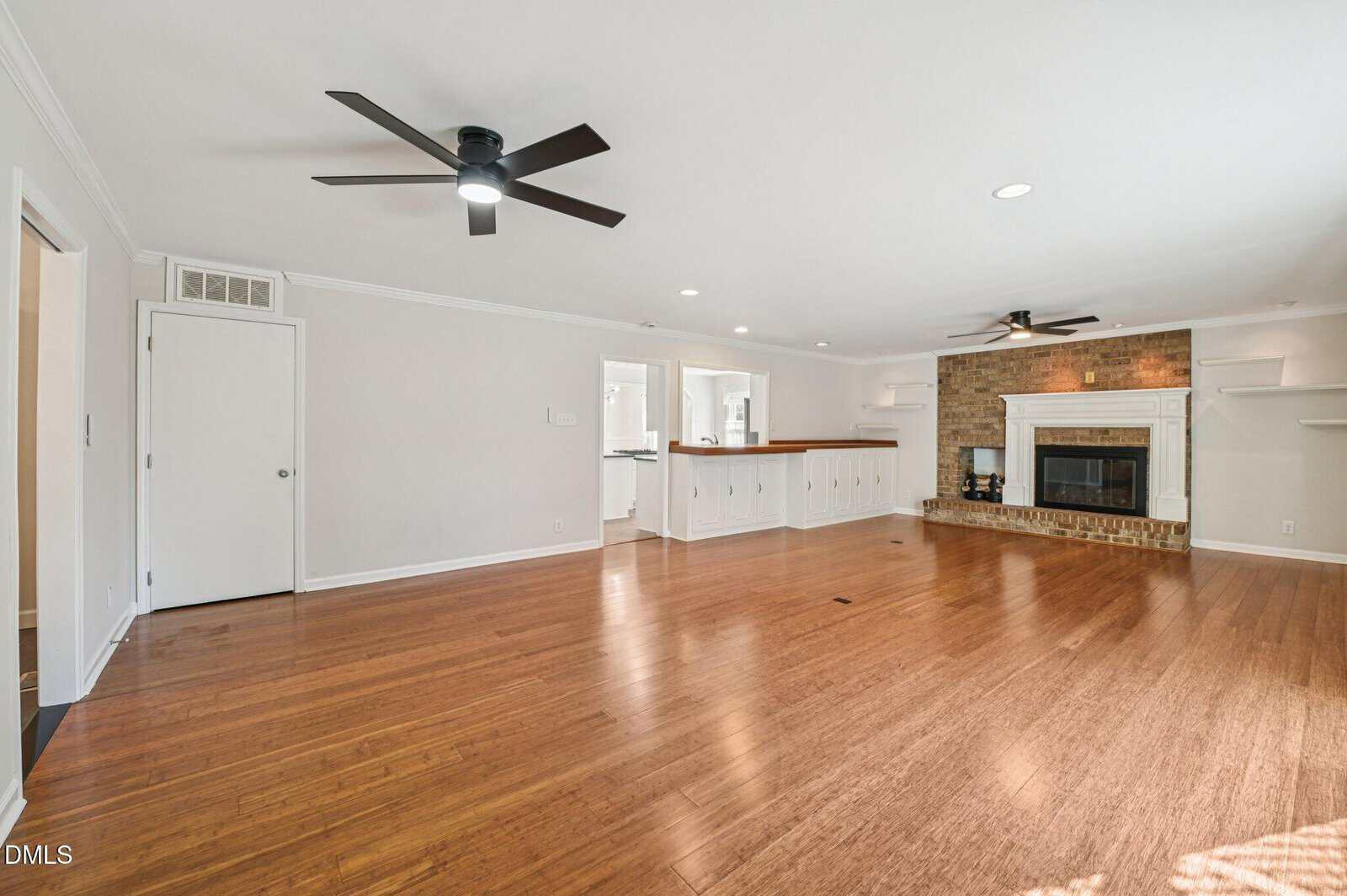 A spacious living room with wood flooring, white walls, a brick fireplace, built-in shelves, ceiling fans, recessed lighting, and a view into the adjacent kitchen area.