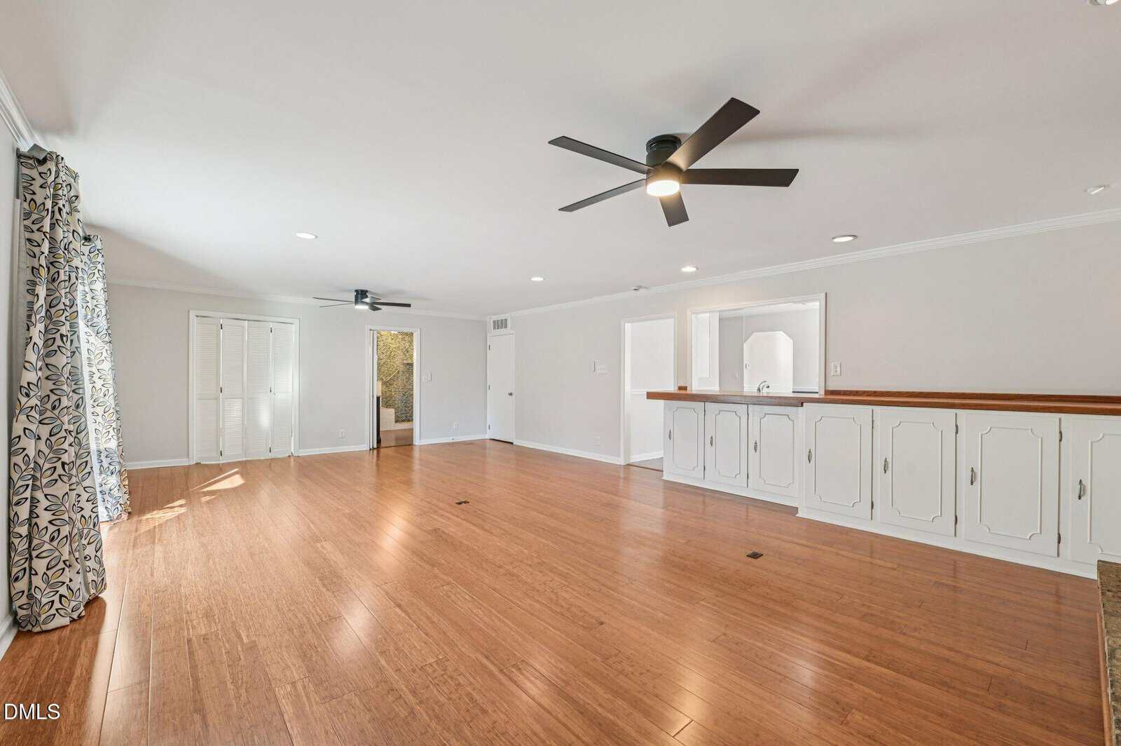Spacious living room with light wood flooring, white walls, two black ceiling fans, built-in cabinets with a wooden countertop, and large windows with patterned curtains letting in natural light.
