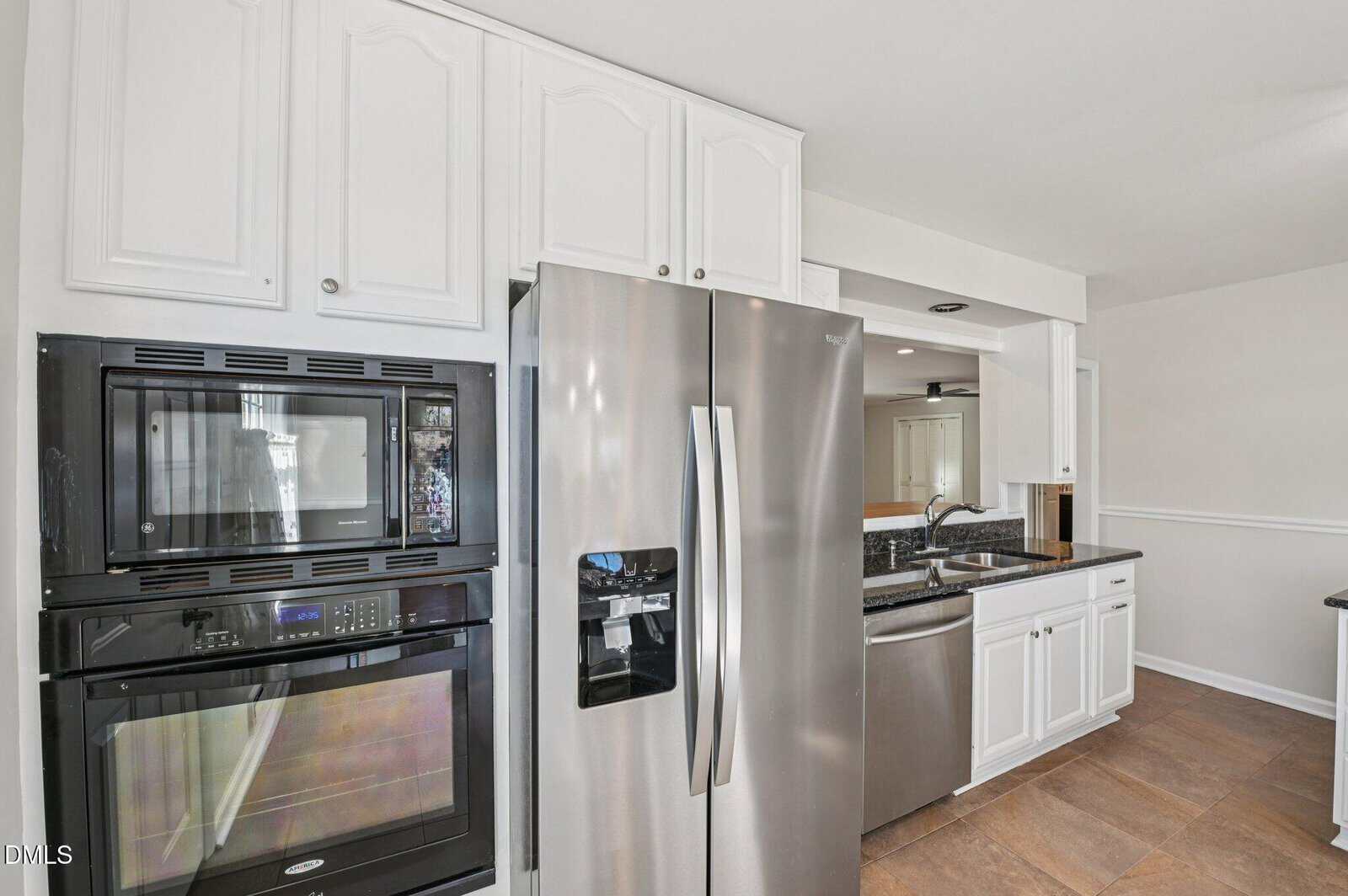 A modern kitchen with white cabinets, stainless steel double-door refrigerator, built-in microwave, and oven, black countertops, double sink, and brown tile floor.