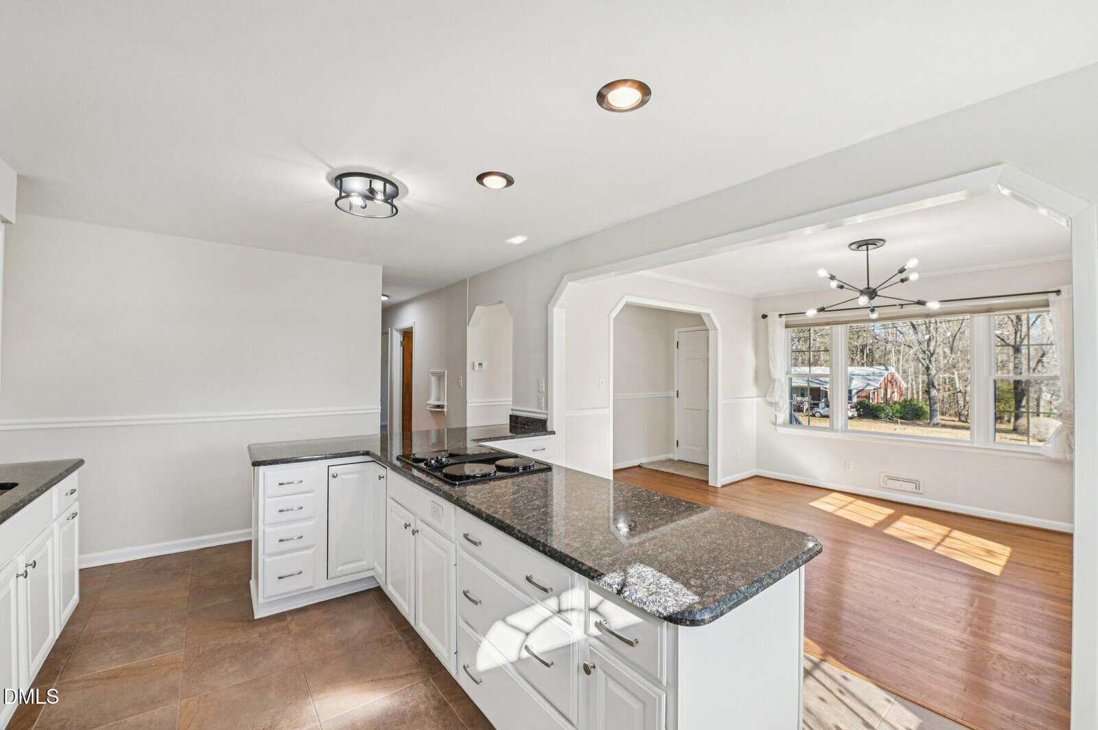 A spacious kitchen with white cabinets, dark granite countertops, and a center stove opens to a bright dining area with large windows, wood floors, and modern light fixtures.