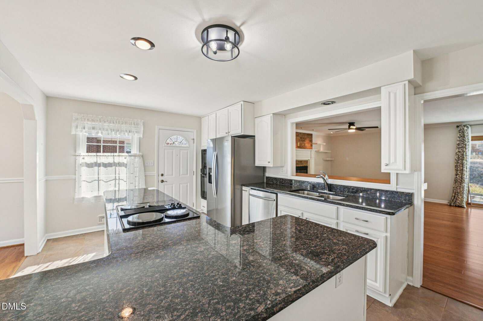 Bright kitchen with black granite countertops, white cabinets, stainless steel appliances, and an open layout. There’s a window over the sink, a ceiling light, and views into the adjoining living area with wood flooring.