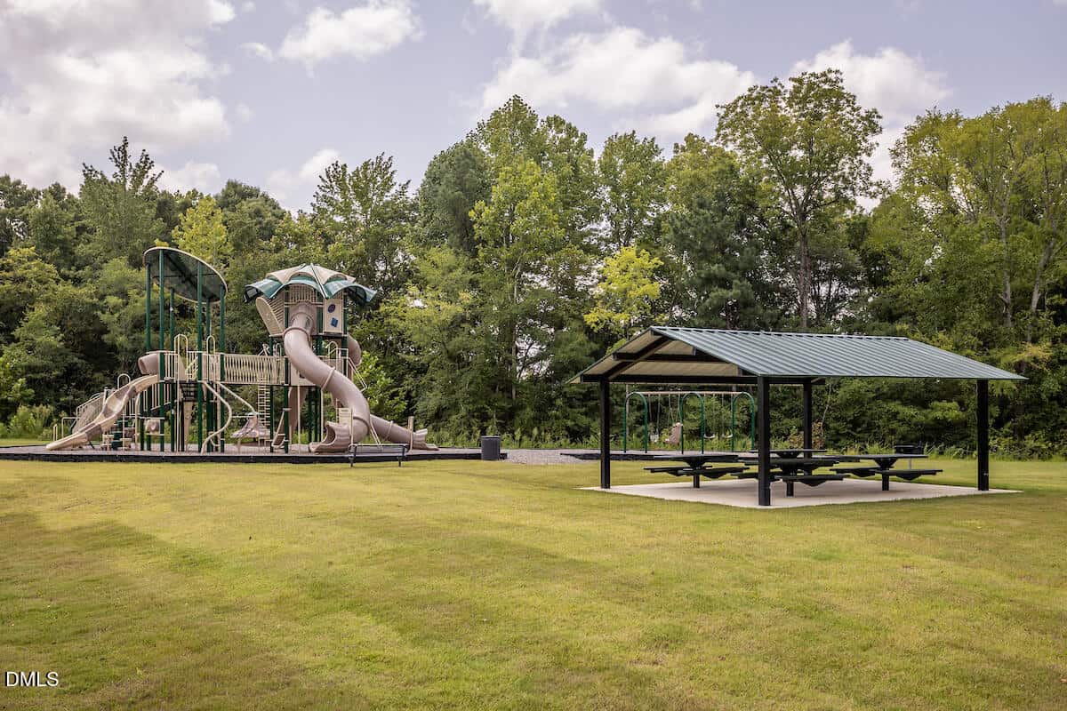 A playground with slides and climbing structures stands next to a covered picnic area with tables on a grassy field, surrounded by trees under a partly cloudy sky.