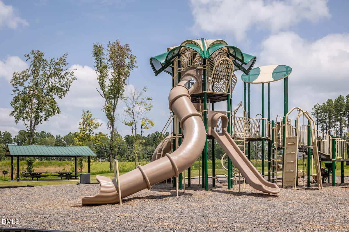 A playground with a large, green and tan play structure featuring multiple slides, climbing areas, and platforms, set on mulch with picnic tables and grassy areas in the background under a partly cloudy sky.