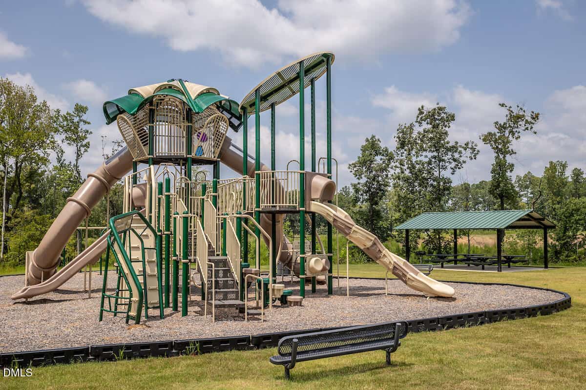 A large playground structure with multiple green and tan slides, climbing elements, and platforms, set on a gravel surface, with a covered picnic area and benches nearby on a grassy field. Trees and a partly cloudy sky in the background.