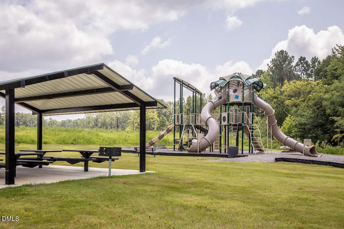 A playground with a large climbing structure and multiple twisting slides stands next to a covered picnic area with tables and a grill, surrounded by green grass and trees under a cloudy sky.