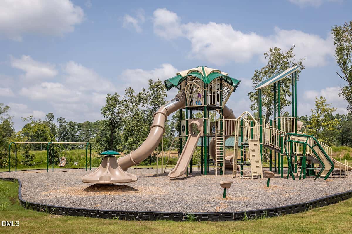 A modern playground with green and beige equipment, including slides, climbing structures, and swings, set on a gravel surface with trees and grass in the background under a partly cloudy sky.