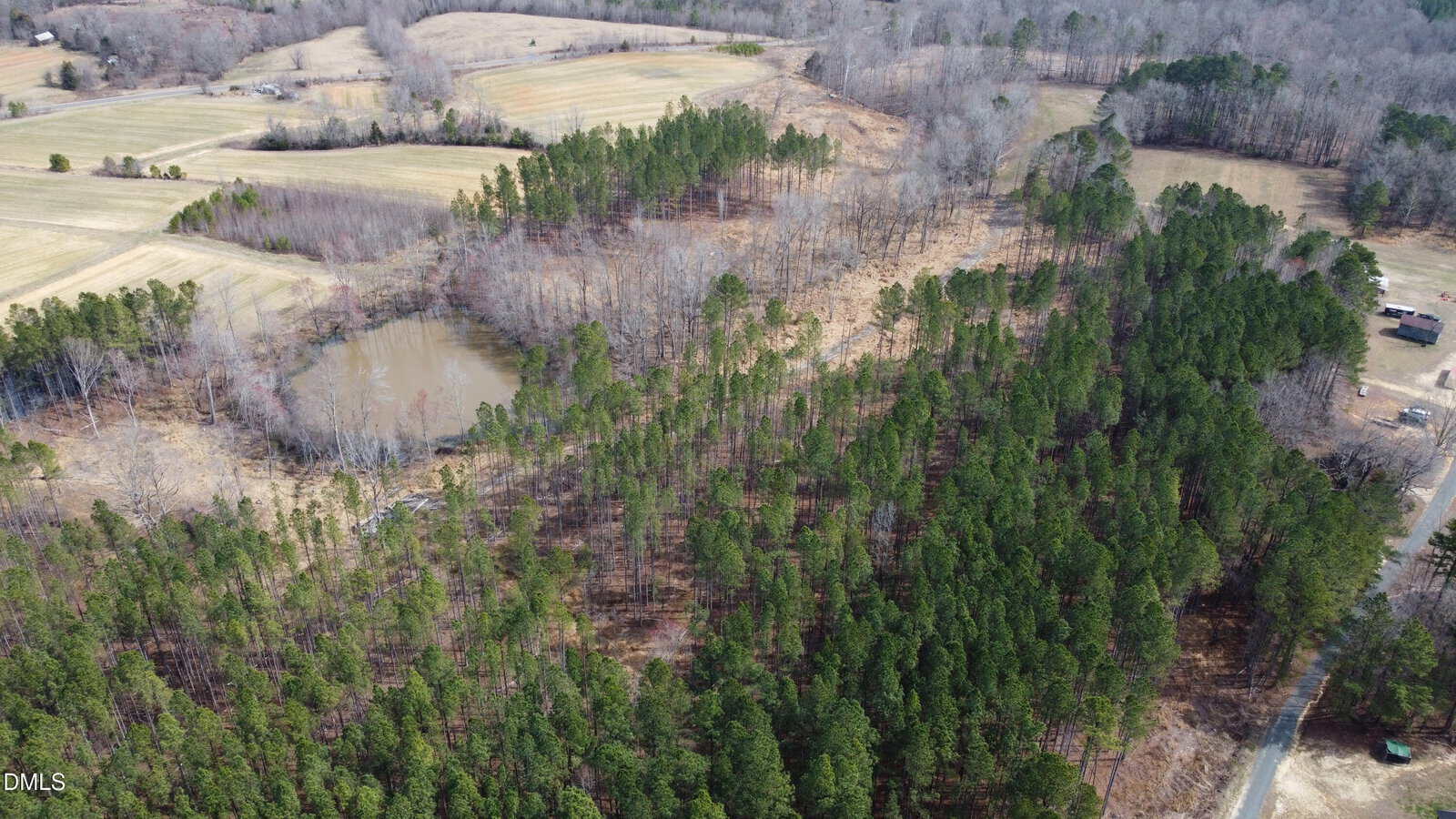 Aerial view of a rural landscape with a forested area, a small pond, open fields, and scattered trees. A road and a few buildings are visible in the distance. The photo appears to be taken in late winter or early spring.