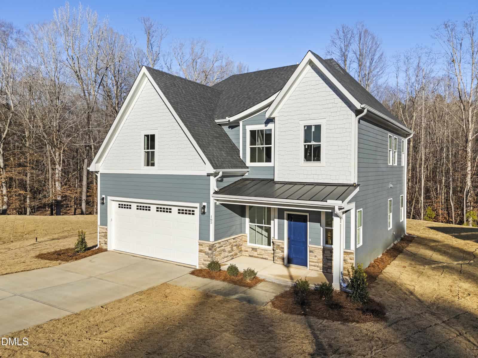 Two-story modern house with light blue siding, white trim, and a dark gray roof. Features a double garage, large windows, and a front porch. The yard is mostly bare with sparse landscaping and surrounded by trees.