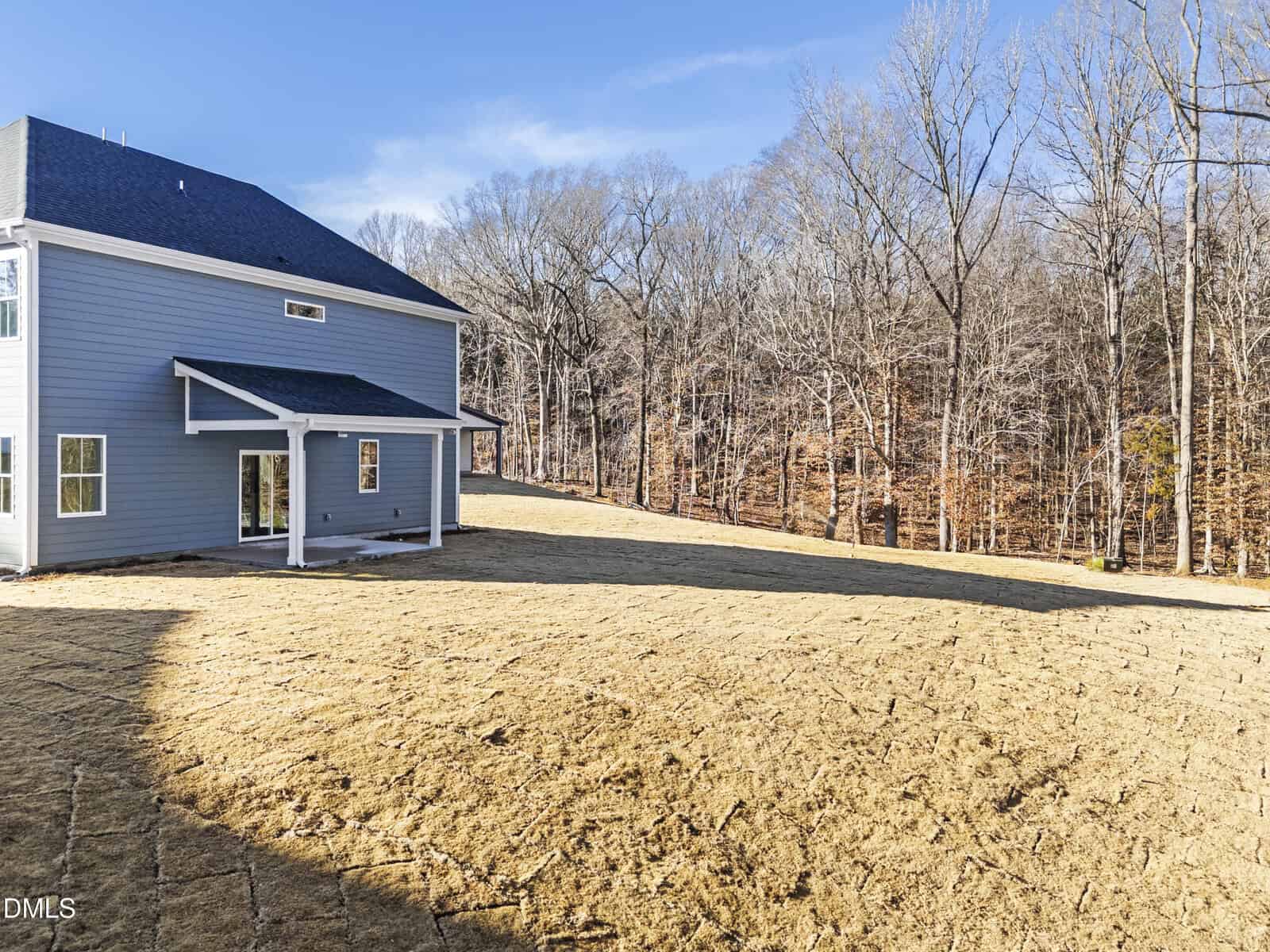 A new two-story house with gray siding and a covered back patio overlooks a spacious, recently seeded yard and a forest of leafless trees under a clear blue sky.