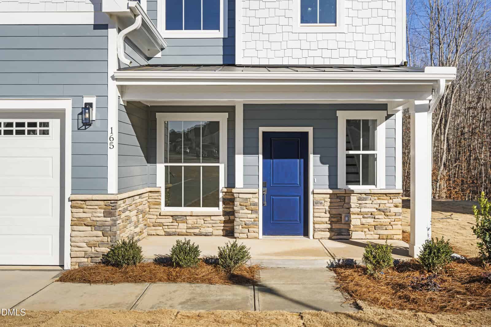 A modern house exterior features blue siding, white trim, stone accents, a bright blue front door, large windows, and small shrubs along the walkway under a covered entryway.