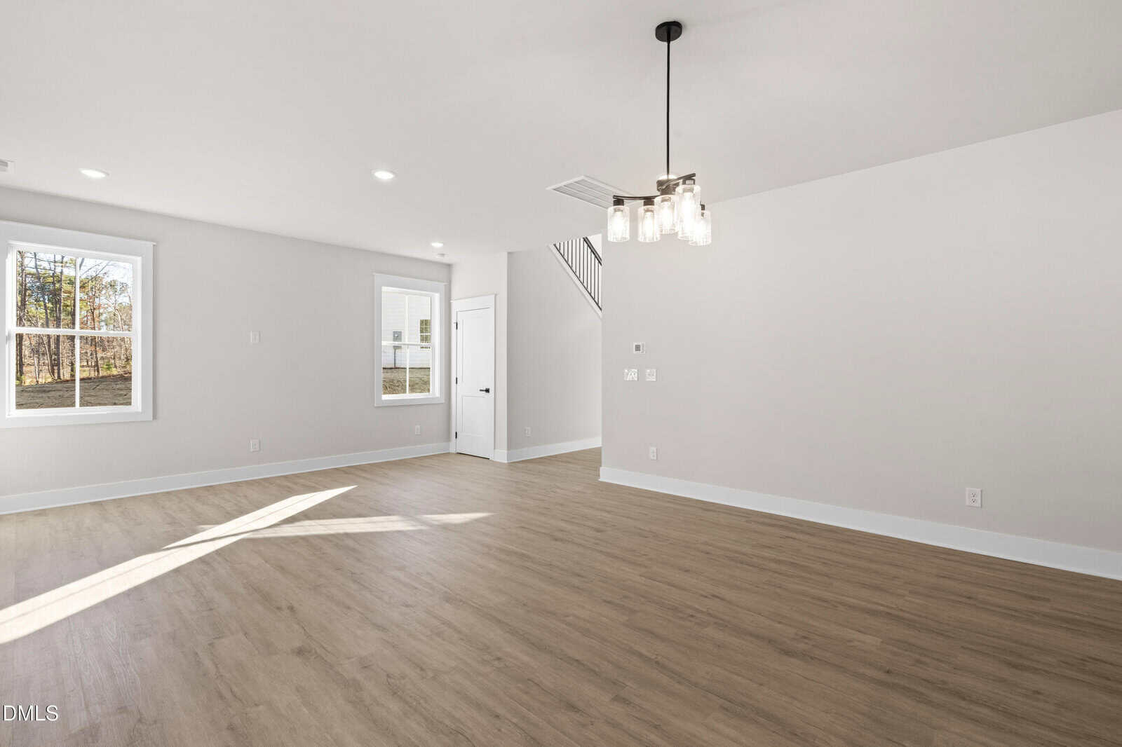 A spacious, empty room with light wood flooring, white walls, a modern ceiling light fixture, two windows, a white door, and stairs leading up. Sunlight streams in through the windows.