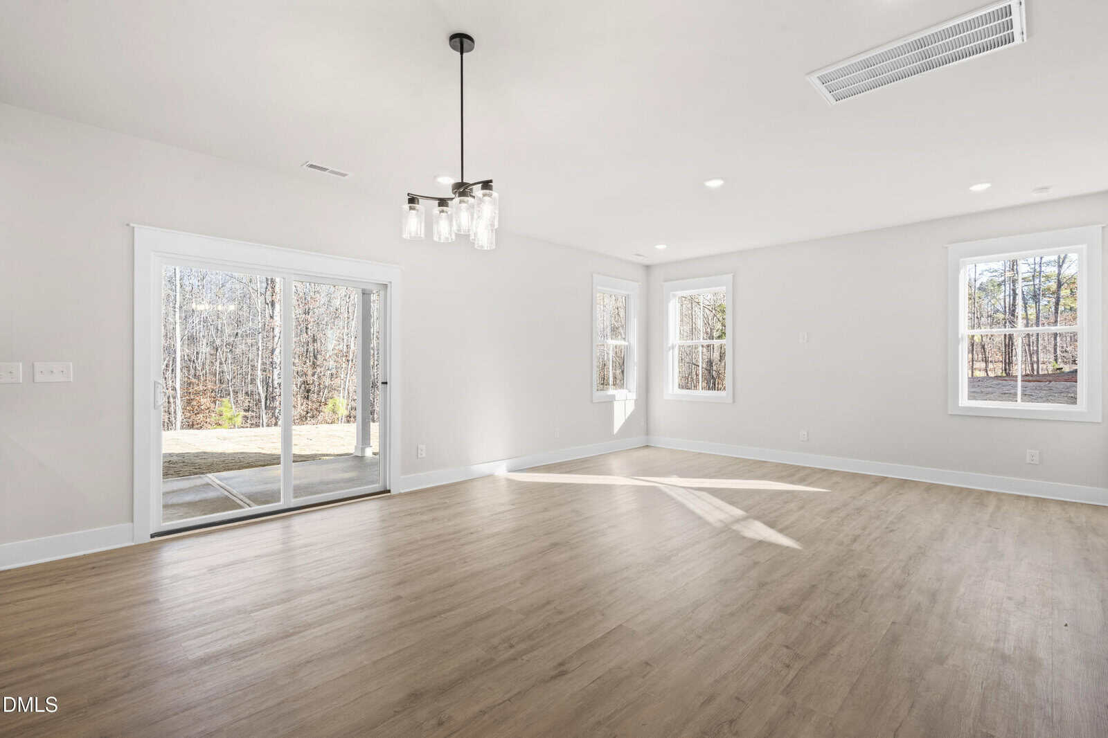 Bright, empty room with light wood flooring, white walls, large windows, a modern ceiling light, and sliding glass doors leading to an outdoor patio with a view of trees.