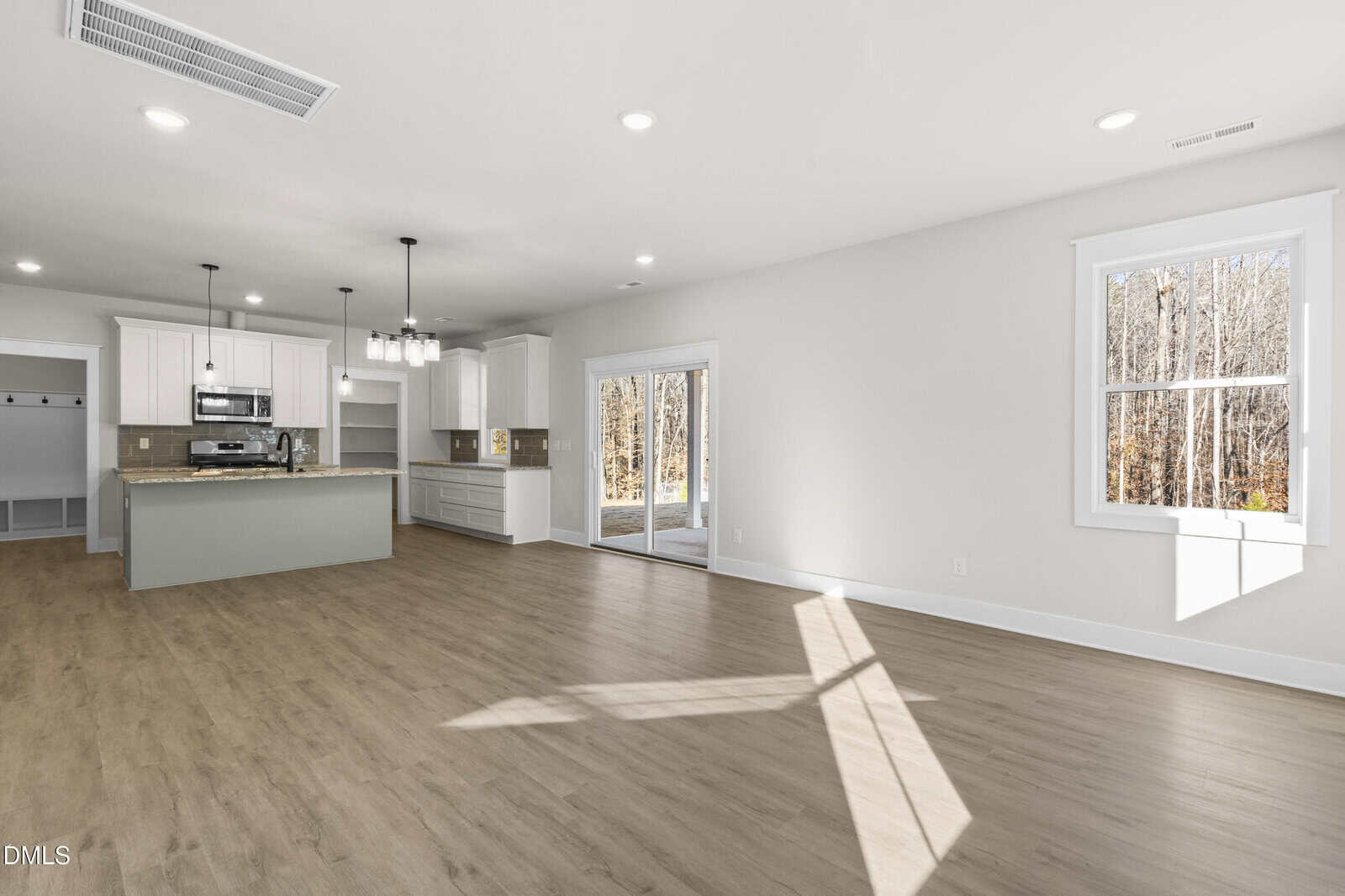 Bright, open-concept living area with wood flooring, white walls, large windows, sliding glass doors, and a modern kitchen featuring an island and pendant lights. Forest view visible through the windows and doors.