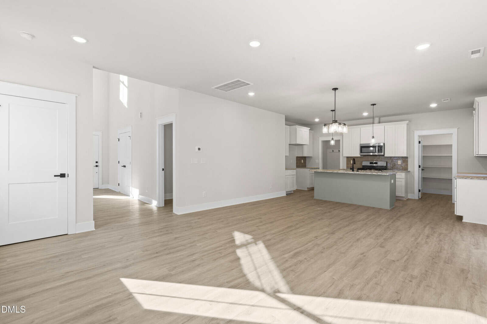 Bright, open-concept kitchen and living area with light wood floors, white walls, large windows casting sunlight, modern pendant lights, stainless steel appliances, and a light green island. Multiple doors lead to other rooms.