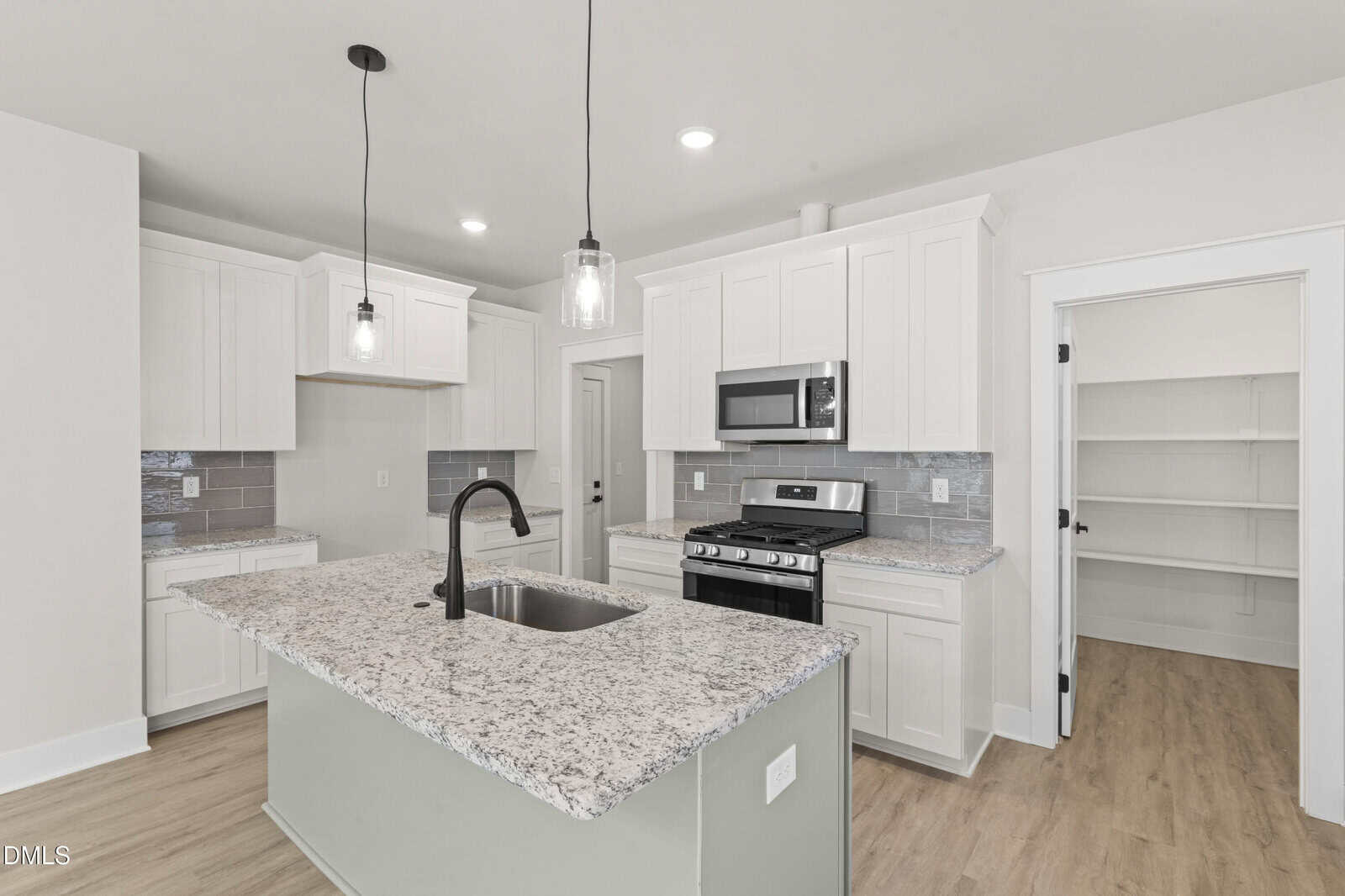 Modern kitchen with white cabinets, granite island with sink, stainless steel appliances, light wood floor, pendant lights, and an open pantry door. Bright, clean, and spacious layout.