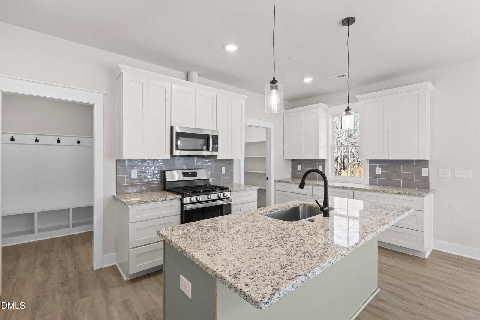 Modern kitchen with white cabinets, granite countertops, stainless steel appliances, gray tile backsplash, island with sink and black faucet, pendant lights, and a view into a mudroom with cubbies.