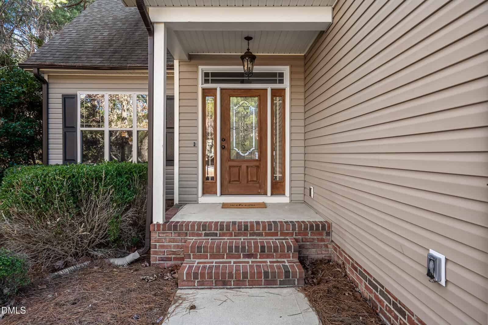 A front porch with brick steps leading to a wooden door with glass panels, surrounded by beige siding. There is a hanging light fixture above the entrance and greenery to the left of the steps.