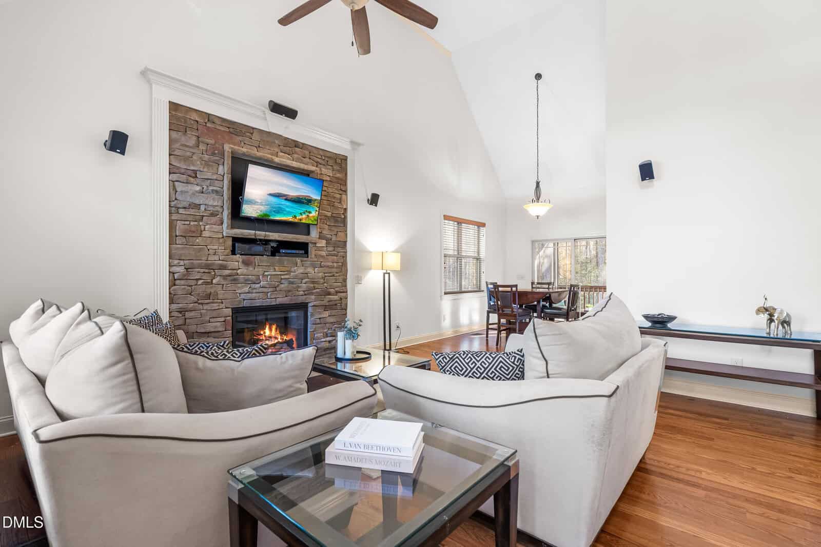 A living room with two beige sofas, a glass side table with books, a stone fireplace with a mounted TV above it, hardwood floors, and an open view into a dining area with large windows.