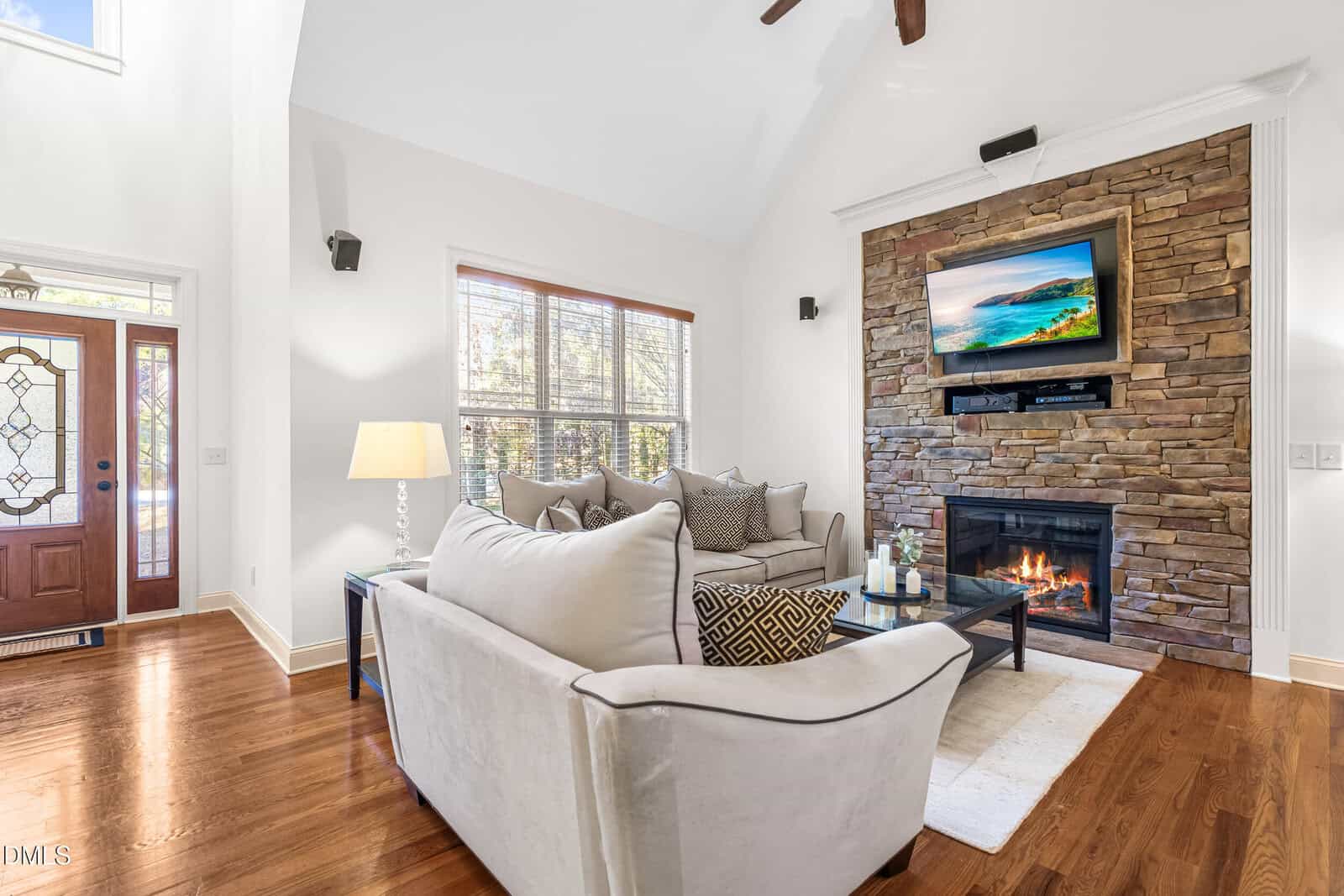 A bright living room with hardwood floors, a stone fireplace with a TV above it, a beige sofa with cushions, a side table with a lamp, large windows, and a decorative glass front door.