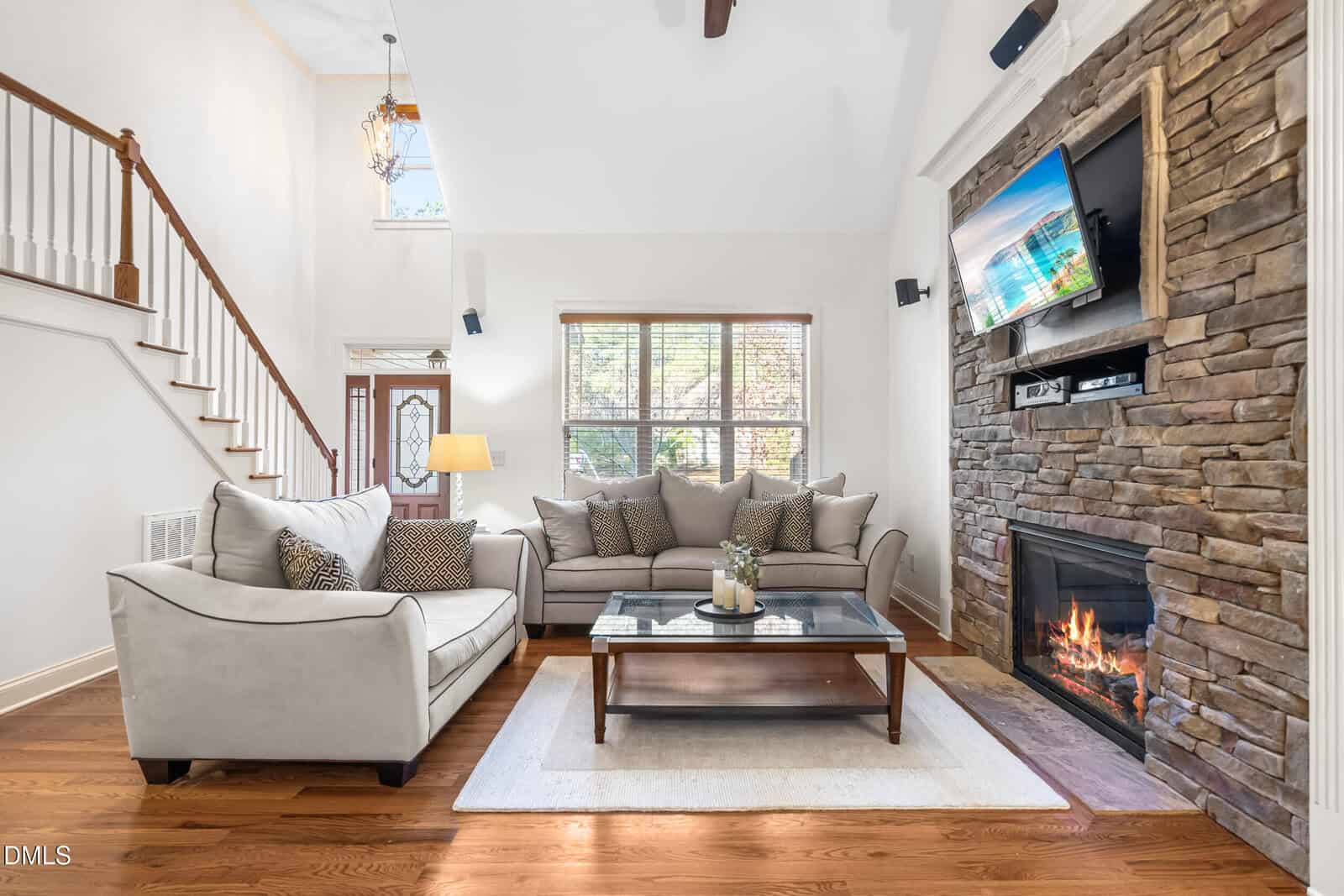 A living room with two light gray sofas, a glass coffee table, a stone fireplace with a TV above it, hardwood floors, and a window letting in natural light. A staircase is visible to the left.