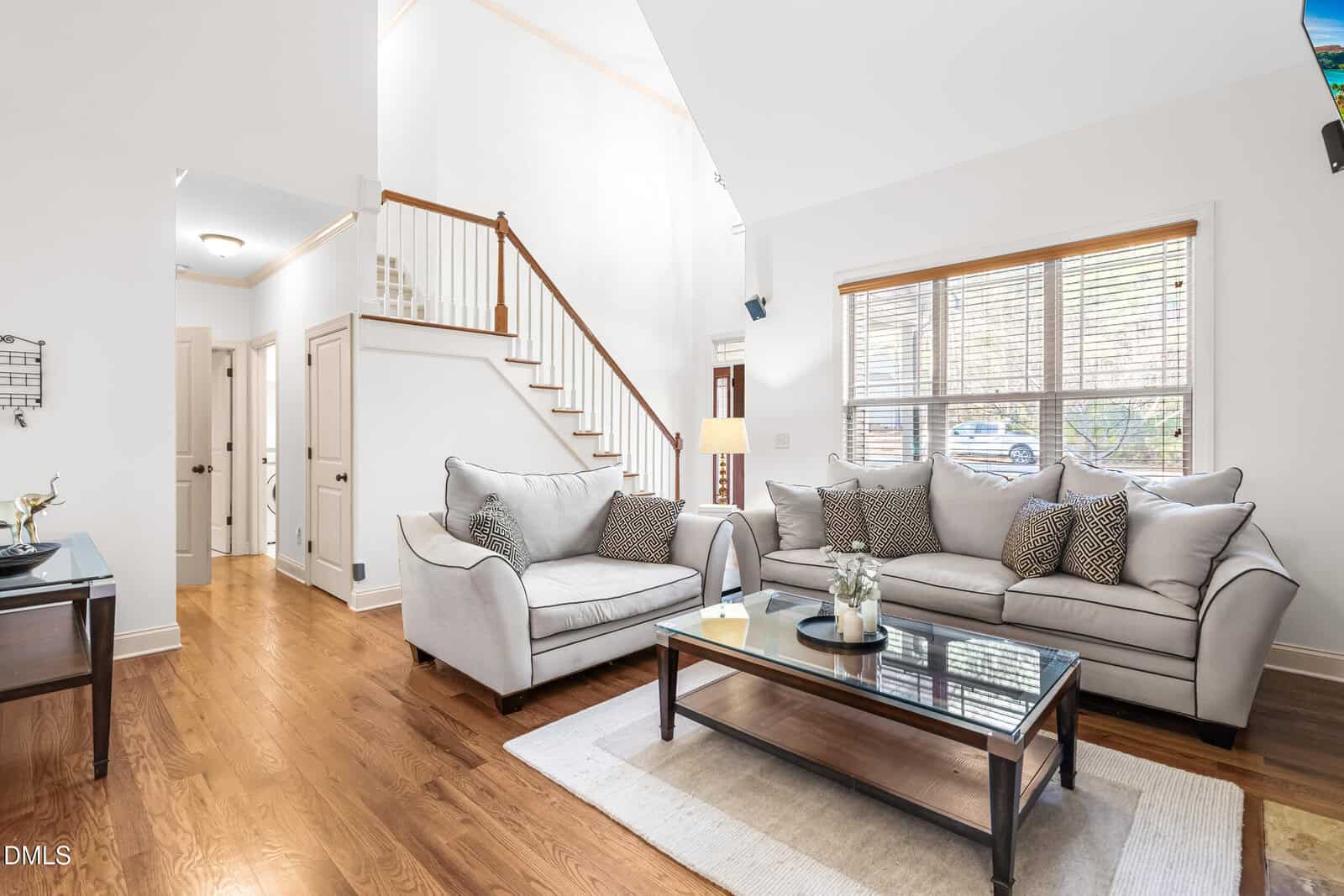A bright living room with hardwood floors, white walls, two light gray sofas with patterned pillows, a glass coffee table, and a staircase leading to the upper floor. Large windows let in natural light.