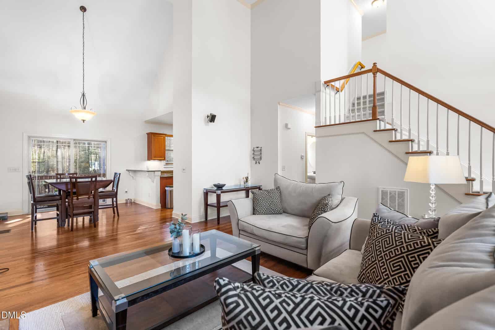 A bright, open living room with hardwood floors, light-colored walls, a gray sofa set, a glass coffee table, and a view into the dining area and kitchen. A staircase with white railings is visible in the background.