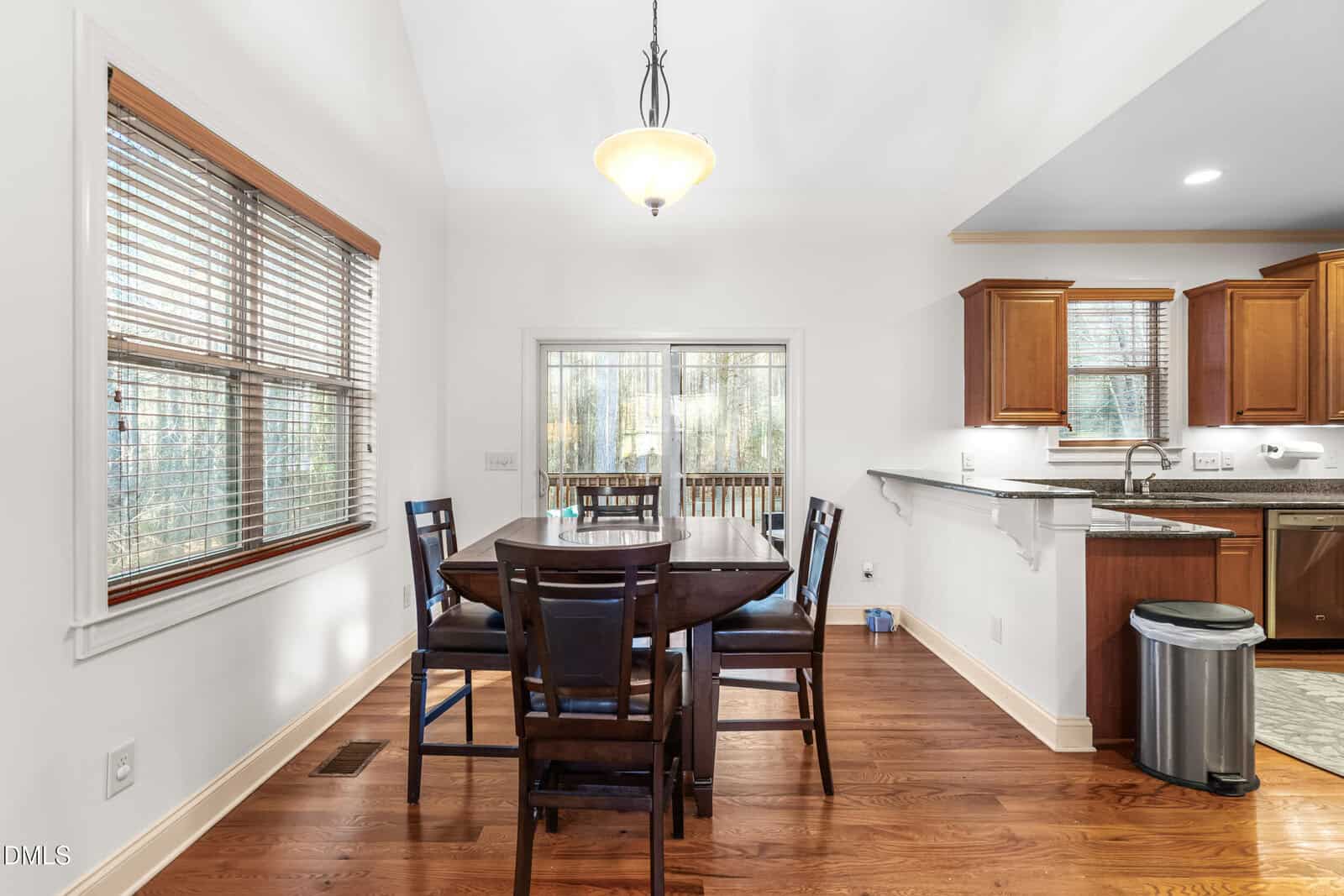 A dining area with a dark wooden table and five chairs on hardwood flooring. Large windows and a glass door provide natural light; adjacent to a kitchen with wooden cabinets and stainless steel appliances.
