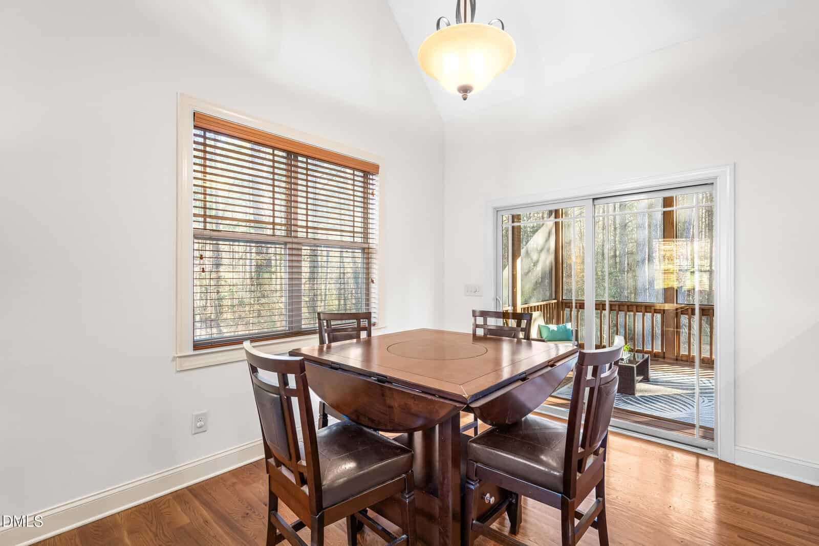 A dining area with a square wooden table and four chairs, wood flooring, a window with blinds, a ceiling light, and sliding glass doors leading to a screened porch.