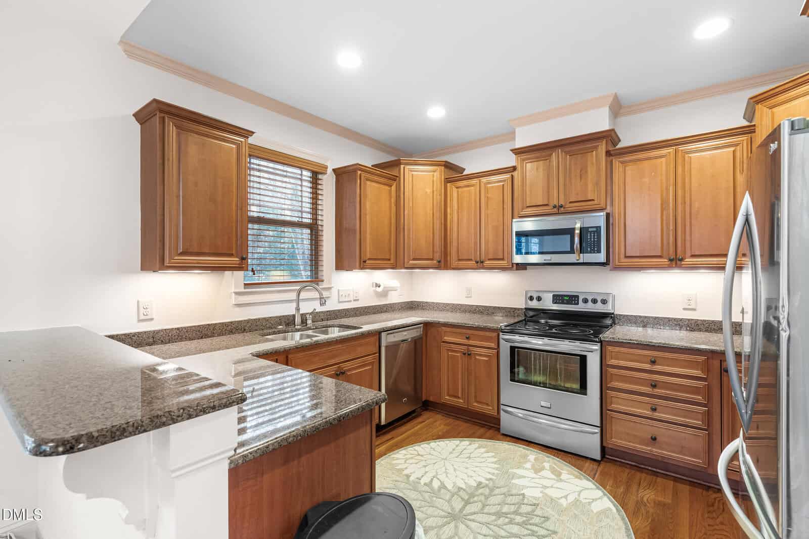 A kitchen with wooden cabinets, granite countertops, stainless steel appliances including an oven, microwave, dishwasher, and refrigerator, a double sink under a window, and a round rug on the floor.