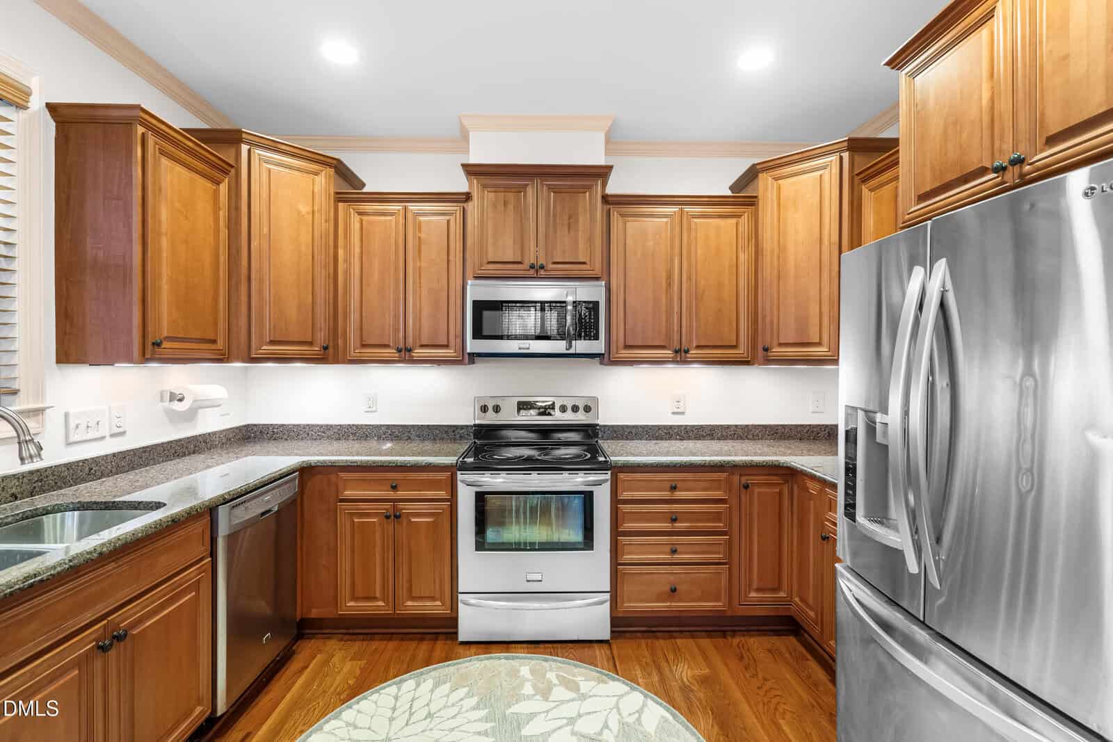 Modern kitchen with wooden cabinets, stainless steel appliances including a refrigerator, oven, microwave, and dishwasher, granite countertops, and hardwood flooring. Bright lighting highlights the clean, organized space.