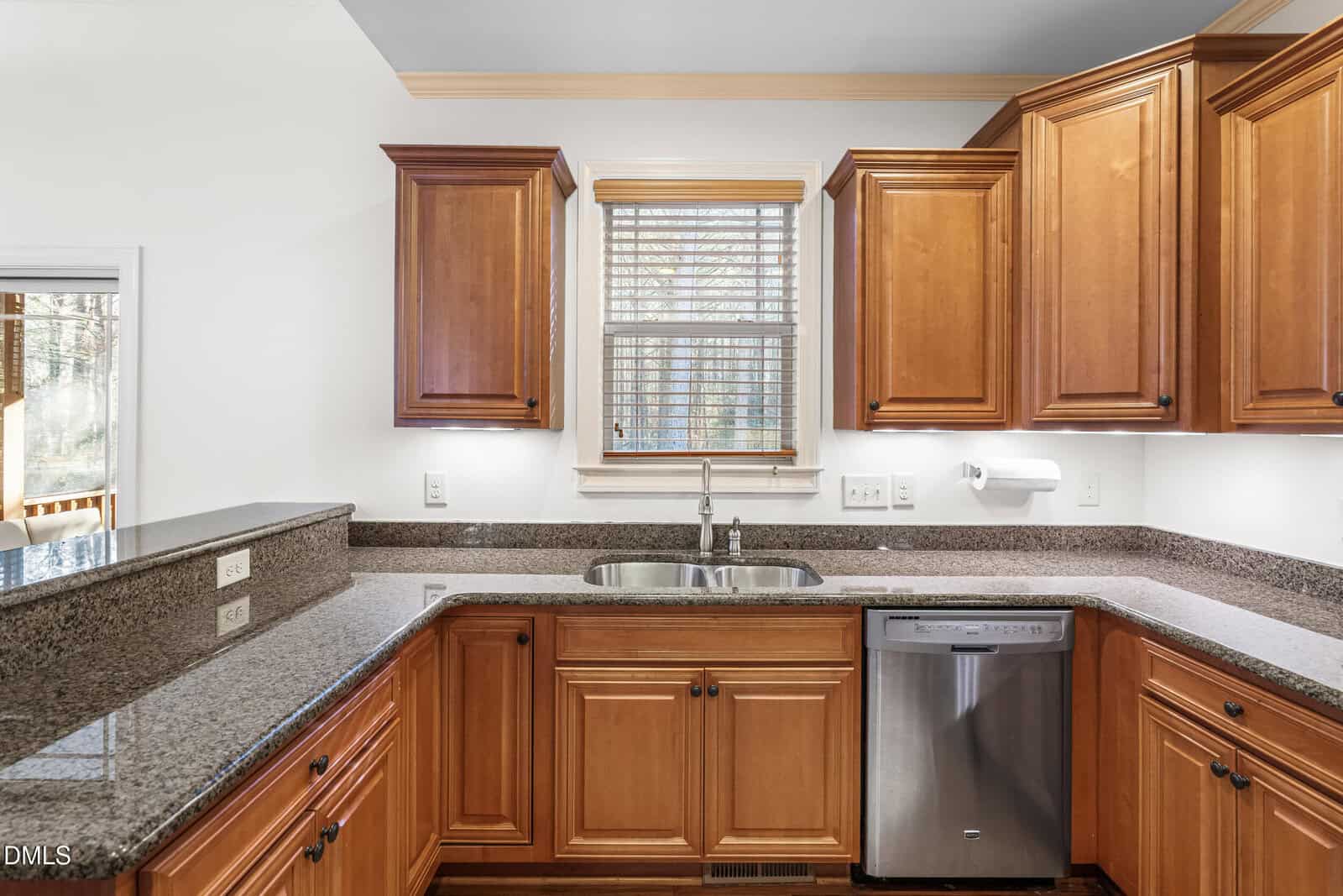 Kitchen with wooden cabinets, granite countertops, a stainless steel dishwasher, a double sink under a window with blinds, and paper towel holder mounted on the wall.
