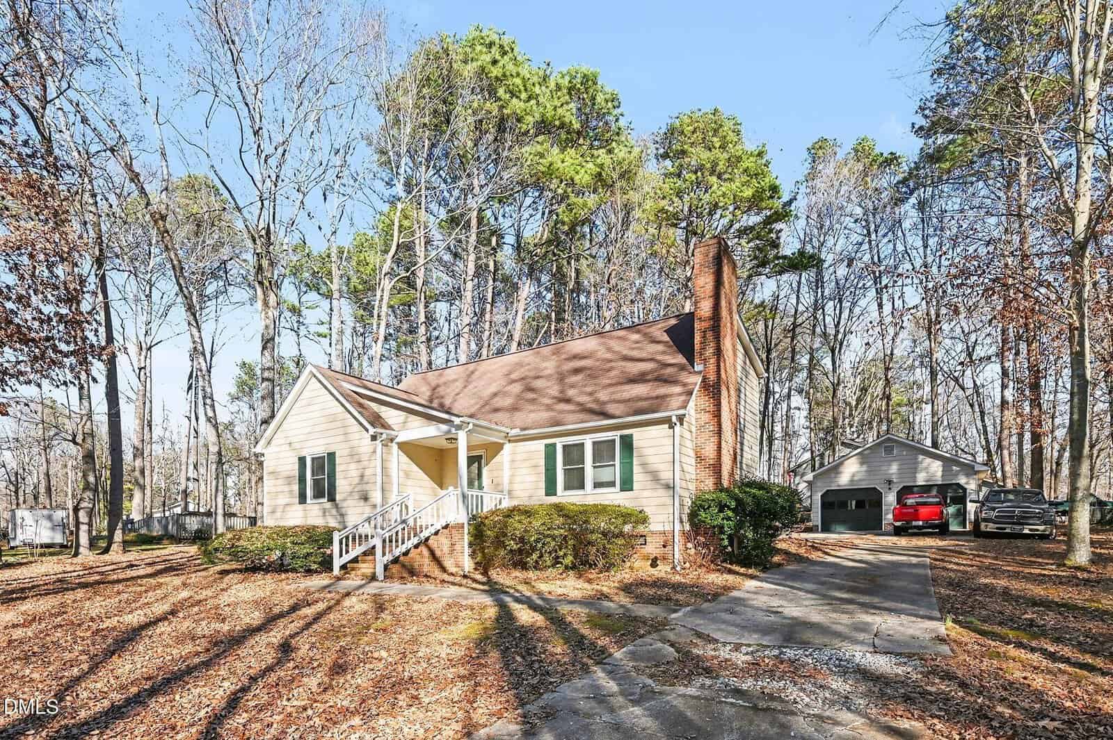 A single-story beige house with a brick chimney and front porch sits among tall trees. A driveway leads to a detached garage, where two vehicles are parked. Fallen leaves cover the ground. It is a sunny day.