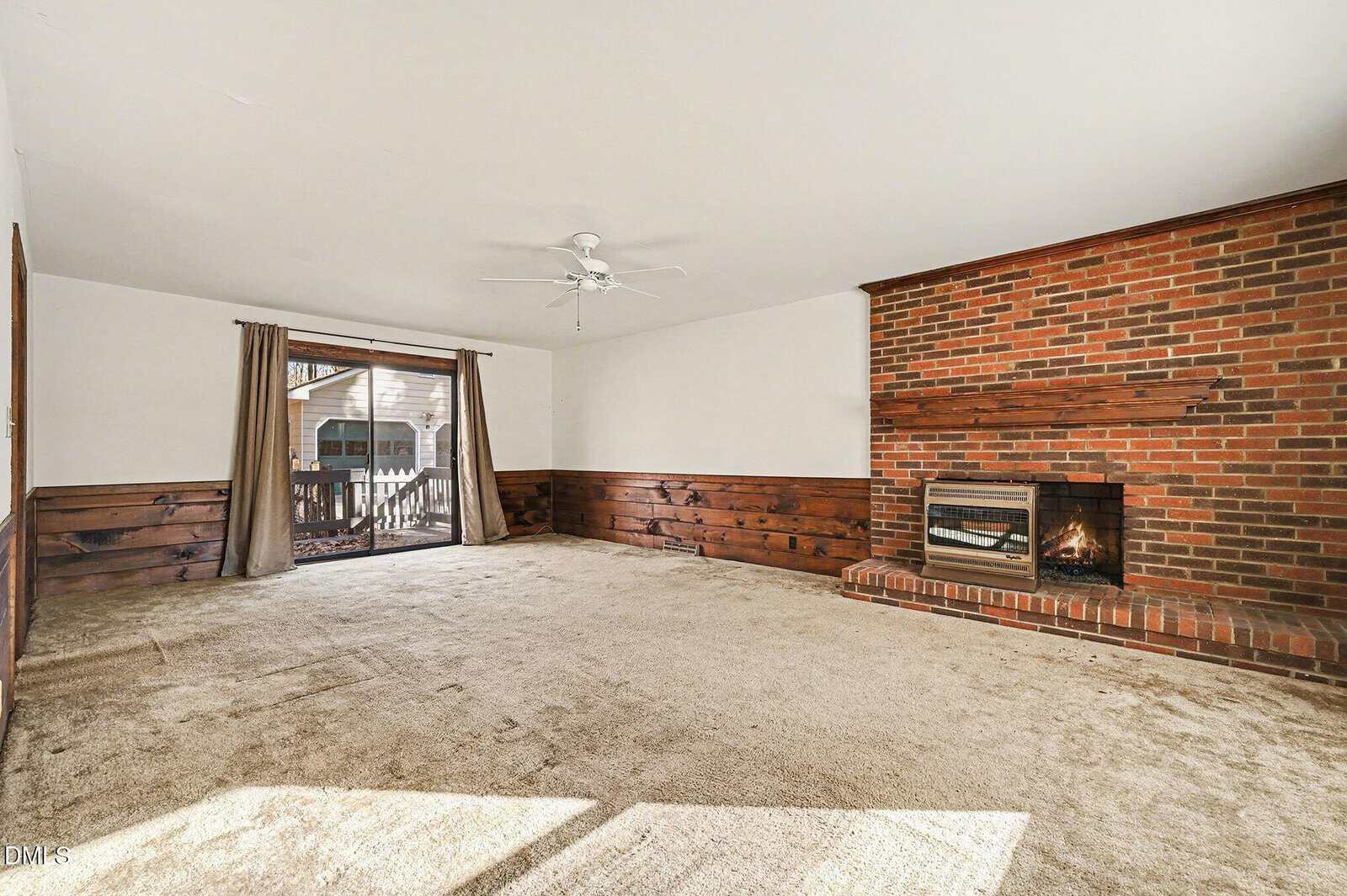 A spacious living room with beige carpet, wood paneling on the lower walls, a ceiling fan, sliding glass doors with curtains, and a brick fireplace with a wood mantel on the right wall.