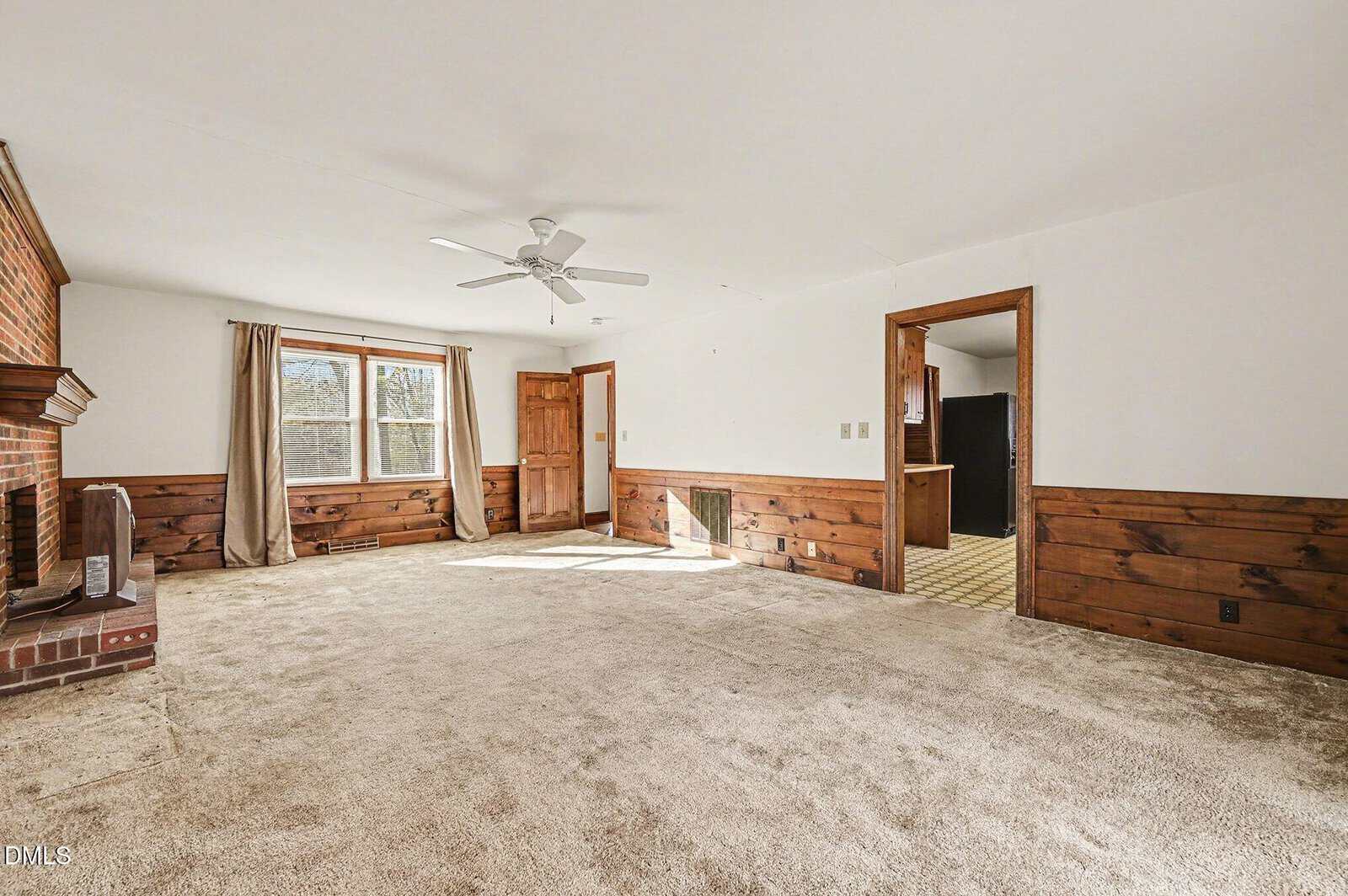 A spacious living room with beige carpet, wood paneling along the lower walls, large windows with curtains, a brick fireplace on the left, a ceiling fan, and two doorways leading to other rooms.