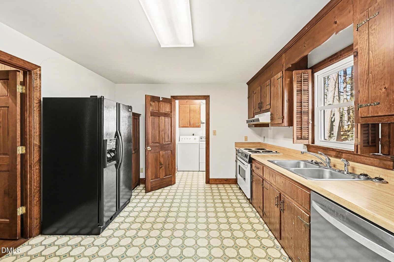 A kitchen with wood cabinets, a double sink, black refrigerator, dishwasher, electric stove, and hood. A door opens to a laundry area with a washer and dryer. The floor has a patterned vinyl covering.