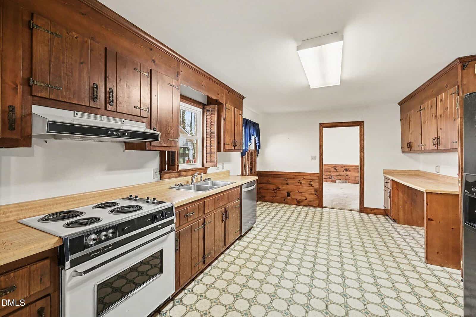 A kitchen with wood cabinets, electric stove, range hood, double sink, laminate countertops, patterned linoleum floor, and fluorescent ceiling light; a doorway leads to an adjacent room.