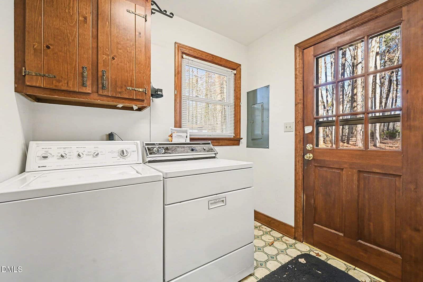 A laundry room with a white washing machine and dryer, wooden cabinets above, a window with blinds, a wooden door with glass panes, and patterned floor tiles. A black mat is placed on the floor near the door.