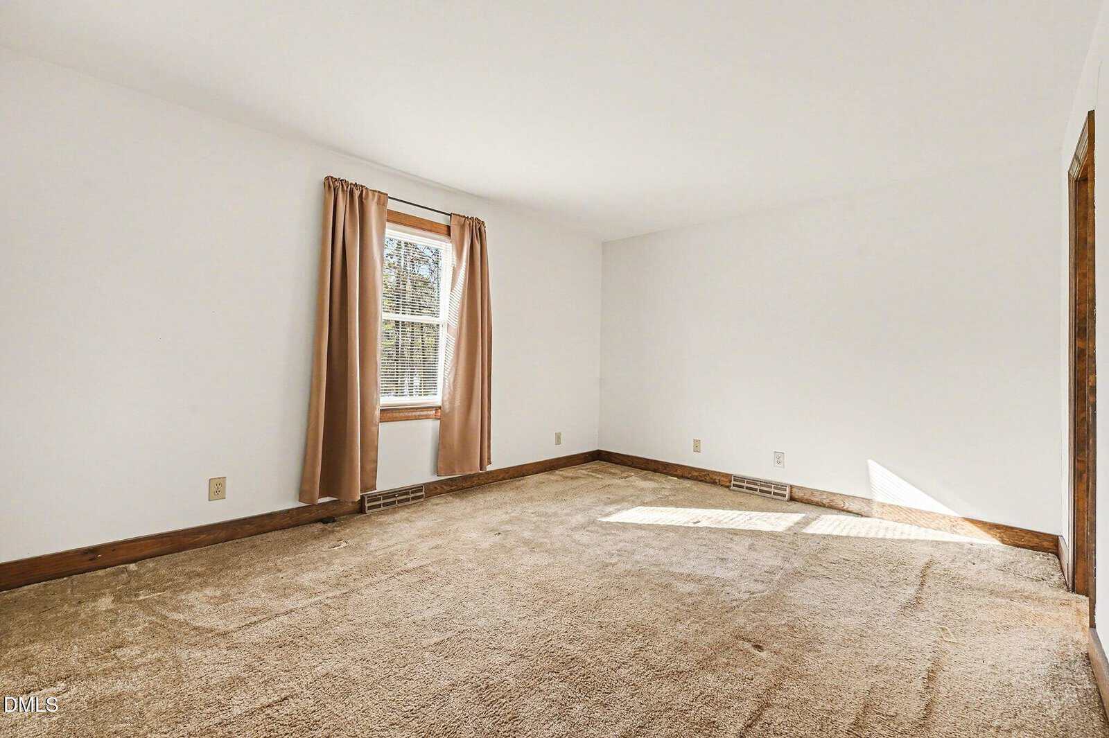 An empty room with beige carpet, white walls, a single window with beige curtains, wood trim, and visible power outlets along the baseboards. Sunlight is streaming in through the window.